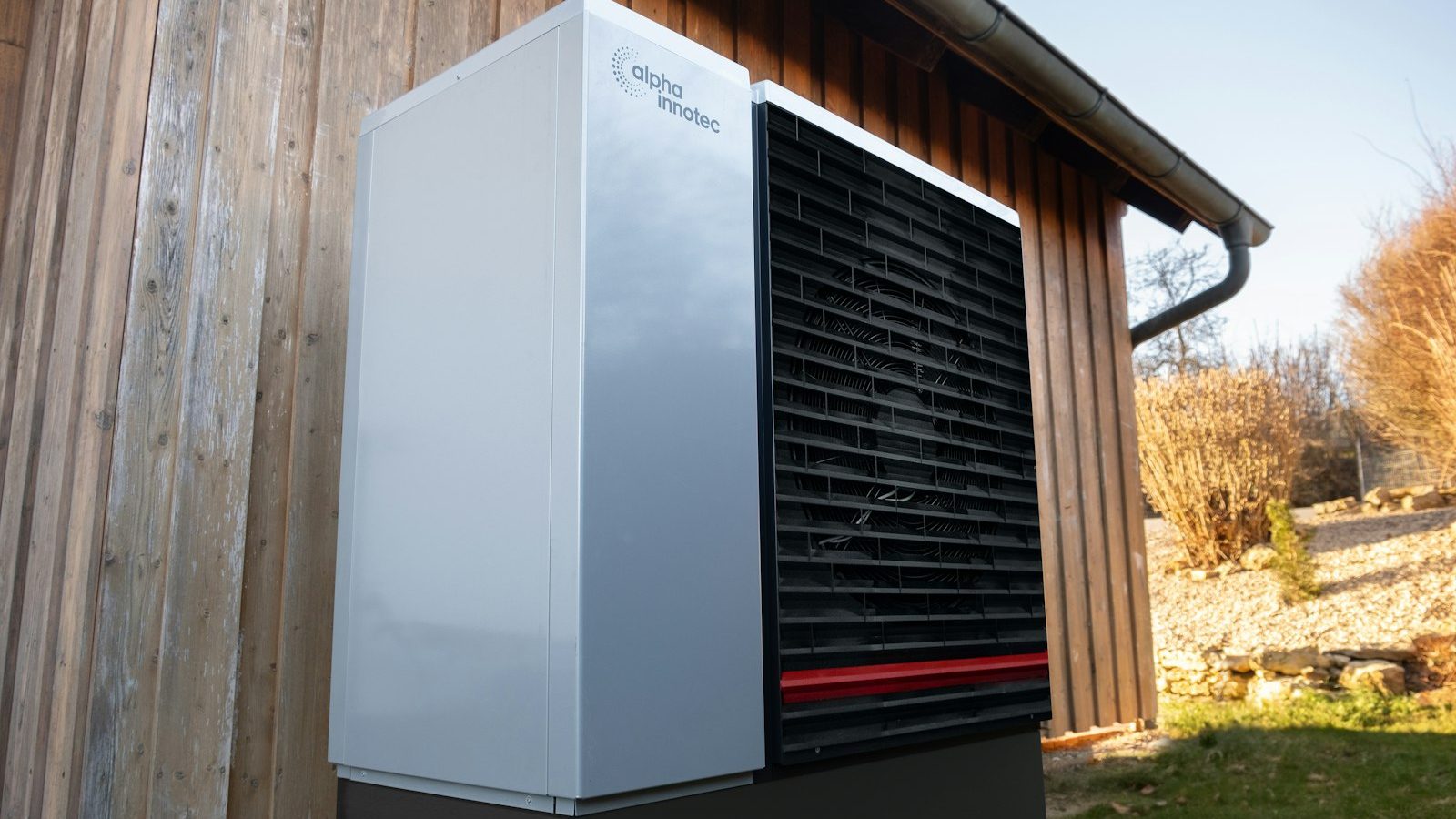 White and black heat pump outdoor unit beside a wood-clad house
