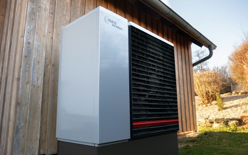 White and black heat pump outdoor unit beside a wood-clad house