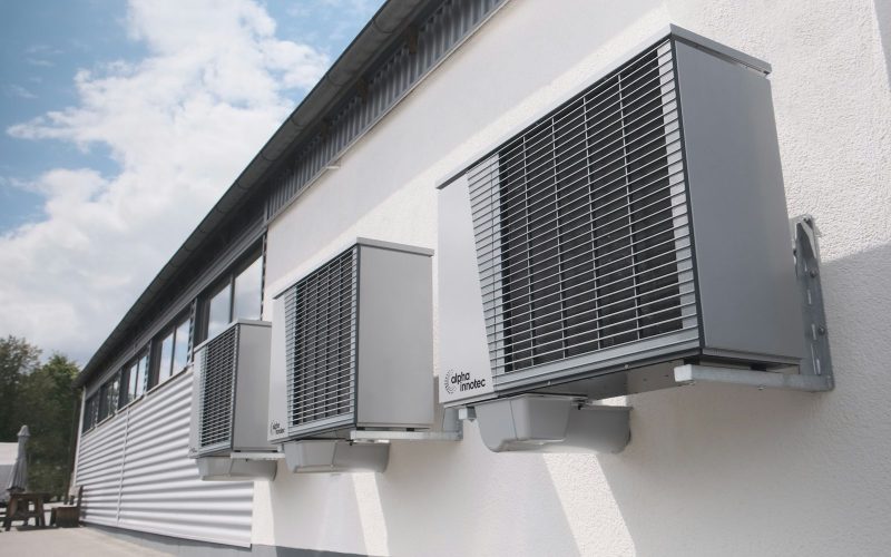 Three gray heat pump outdoor units mounted on a white wall
